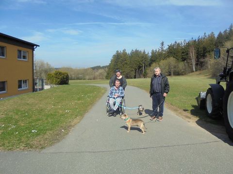 Das Bild zeigt einen Bewohner im Rollstuhl und zwei Angehörige mit zwei Hunden an der Leine