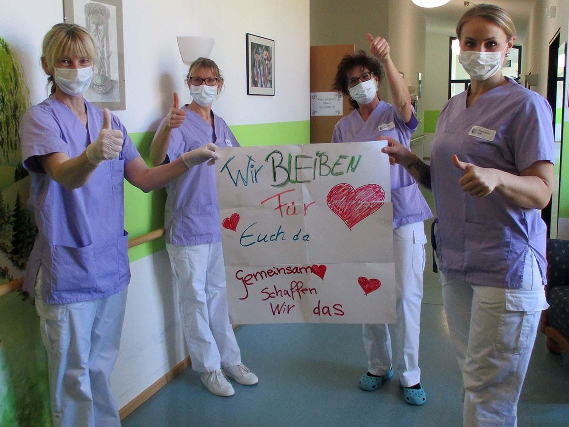 Das Bild zeigt 4 Pflegefachkräfte mit einem aufmunteren Banner gegen den Coronavirus